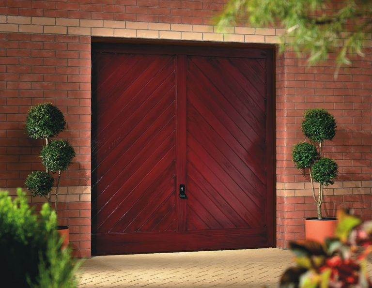 Red wooden garage door with diagonal panel design, framed by brickwork and potted topiary trees – Cristal Windows, Doors & Conservatories