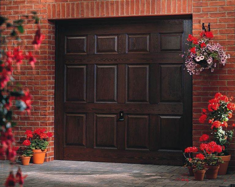 Traditional dark wood-effect panelled up and over garage door framed by red brickwork and surrounded by potted flowers – Cristal Windows, Doors & Conservatories