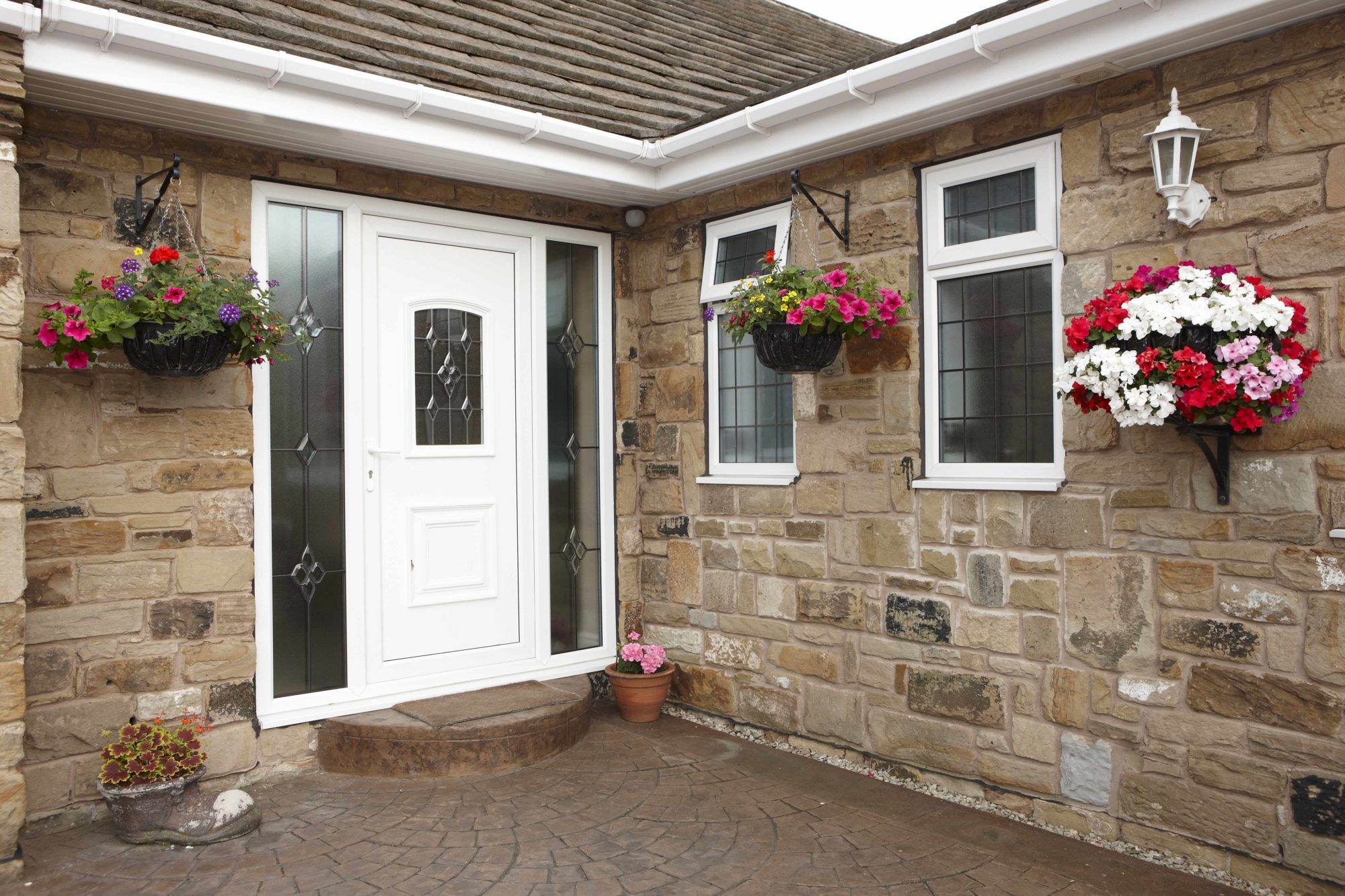 White uPVC residential door with decorative glass panels and matching side panels, set in a stone exterior with hanging flower baskets – Cristal Windows, Doors & Conservatories