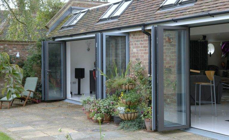 Exterior view of home with multiple open aluminium bi-fold doors in black frame leading to patio and garden – Cristal Windows, Doors & Conservatories