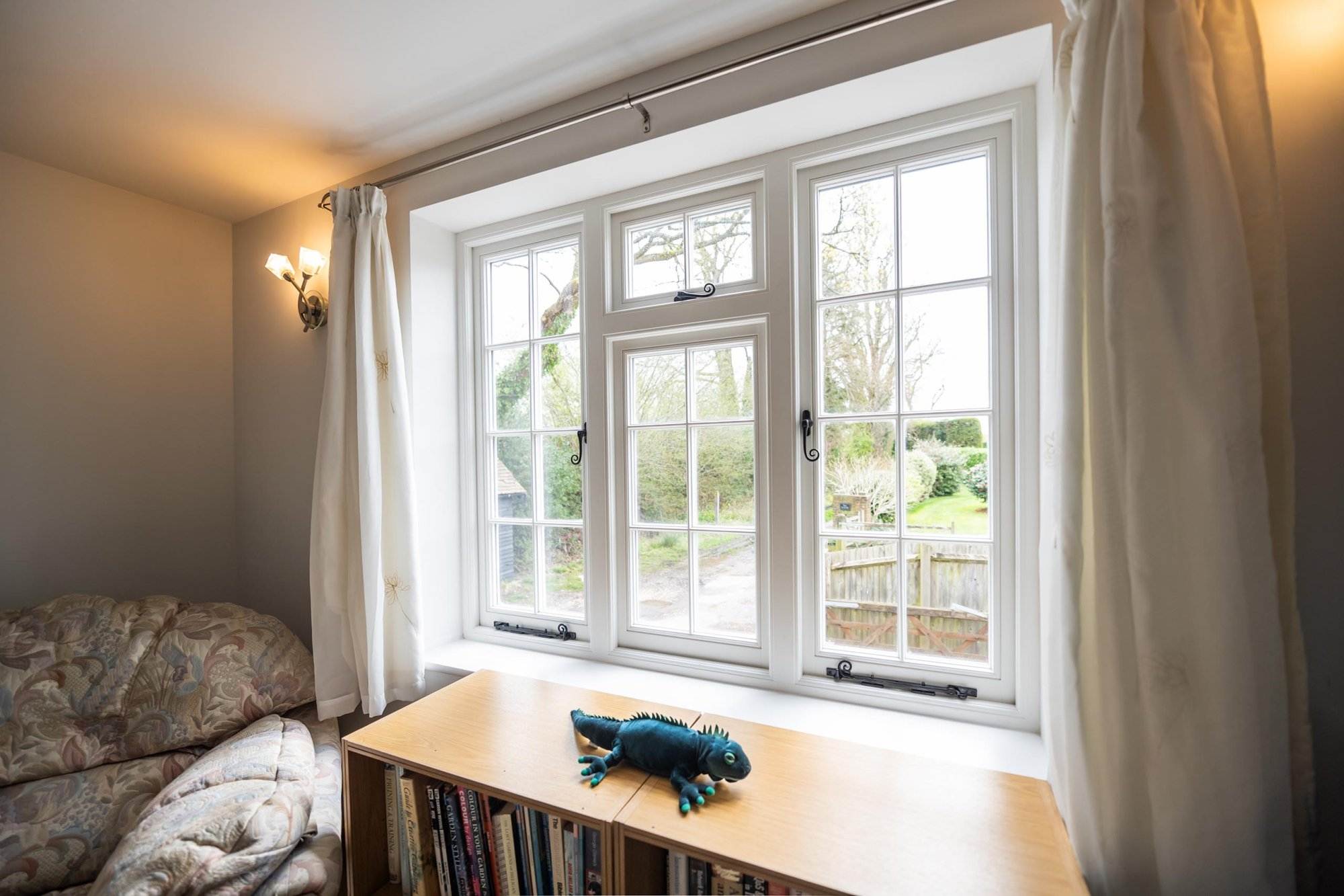 White timber casement window with decorative grid design, overlooking a green garden, shown closed with curtains and bookshelf inside — Cristal Windows, Doors & Conservatories